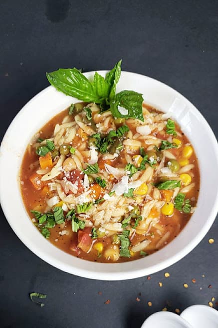 A soup bowl filled with vegan orzo soup and garnished with cheese, herbs and black pepper.