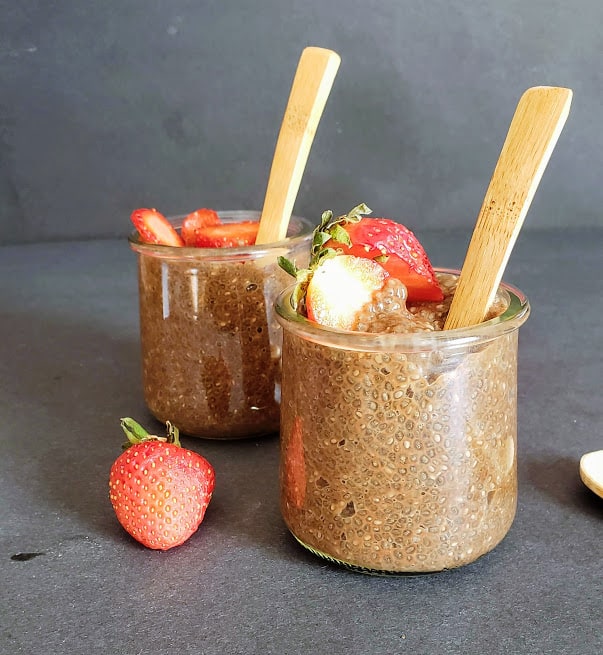 Two glass cups filled with chocolate chia seeds pudding served with fresh strawberries and wooden spoons for eating.