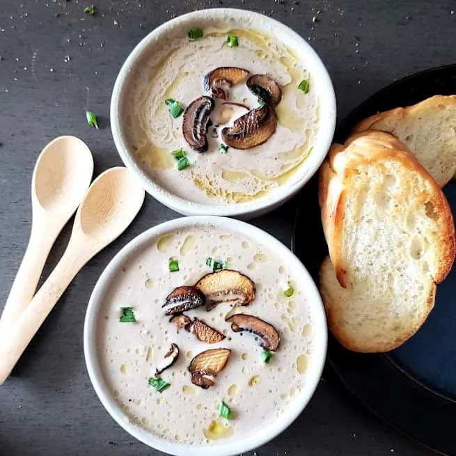 Creamy Mushroom Soup served in two soup bowls. There is toasted bread and wooden spoons on the side. This vegan and gluten free soup is homemade.
