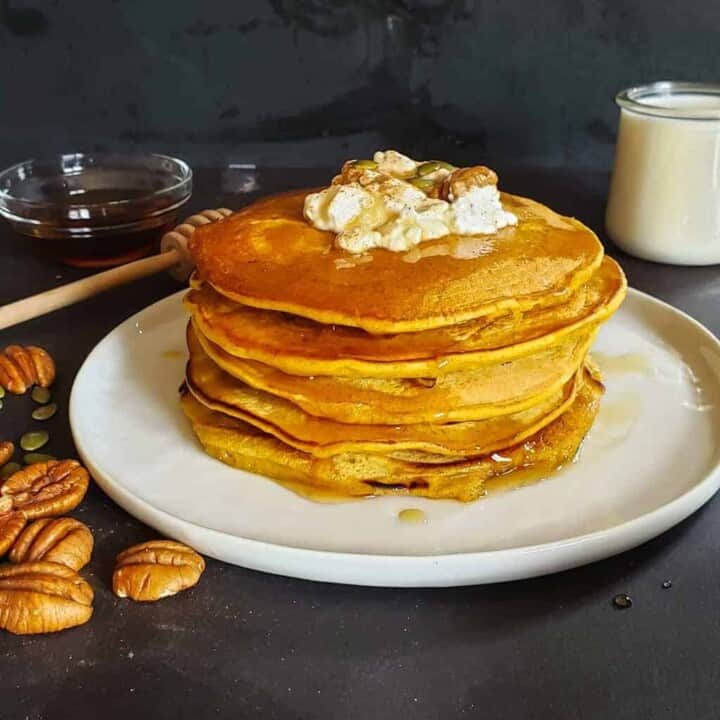 Stack of healthy vegan pumpkin pancakes served with coconut cream, maple syrup and pecans.