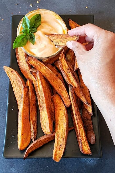 A hand holding a sweet potato fry and dipping it in the sauce.