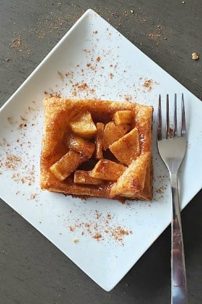 An apple pie puff pastry served with a fork on the white dessert plate.