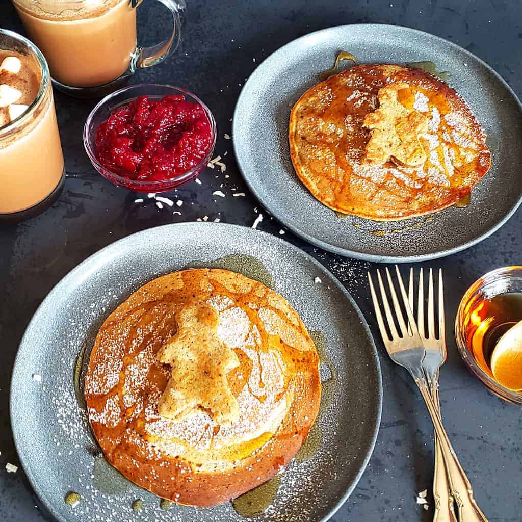 Two plates with vegan gingerbread pancakes along with hot cocoa and cranberry sauce and two forks for eating.