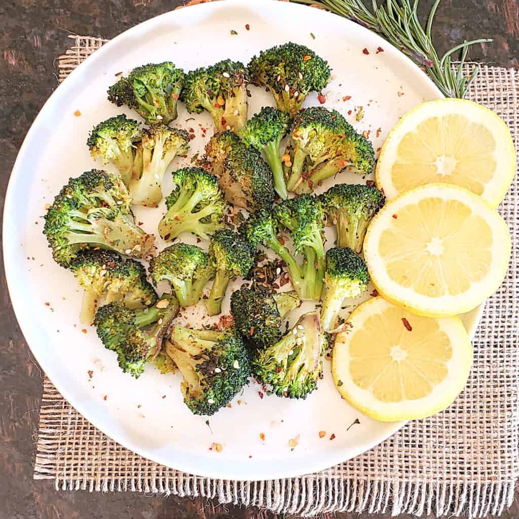 Crispy air fryer broccoli florets garnishes with lemon wedges and rosemary served on a white dinner plate.