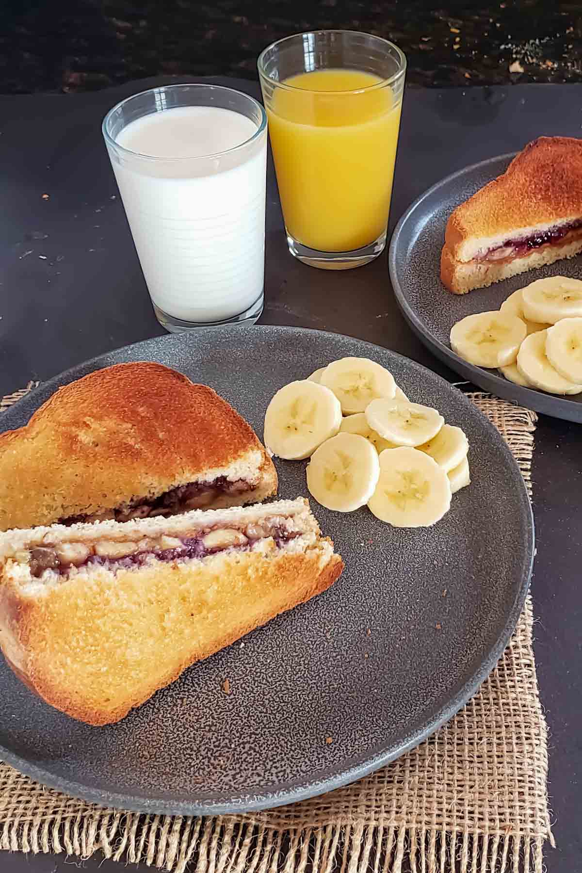 Two plates of pb and j toast sandwich served with cut up banana slices and milk and orange juice.