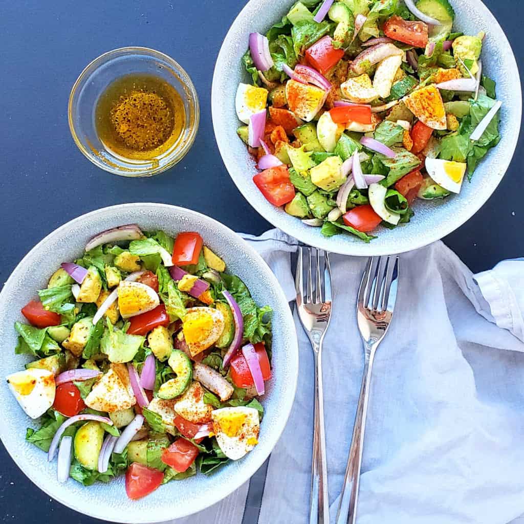 Two salad bowls filled with avocado egg cucumber salad along with two forks on the dinner napkin.