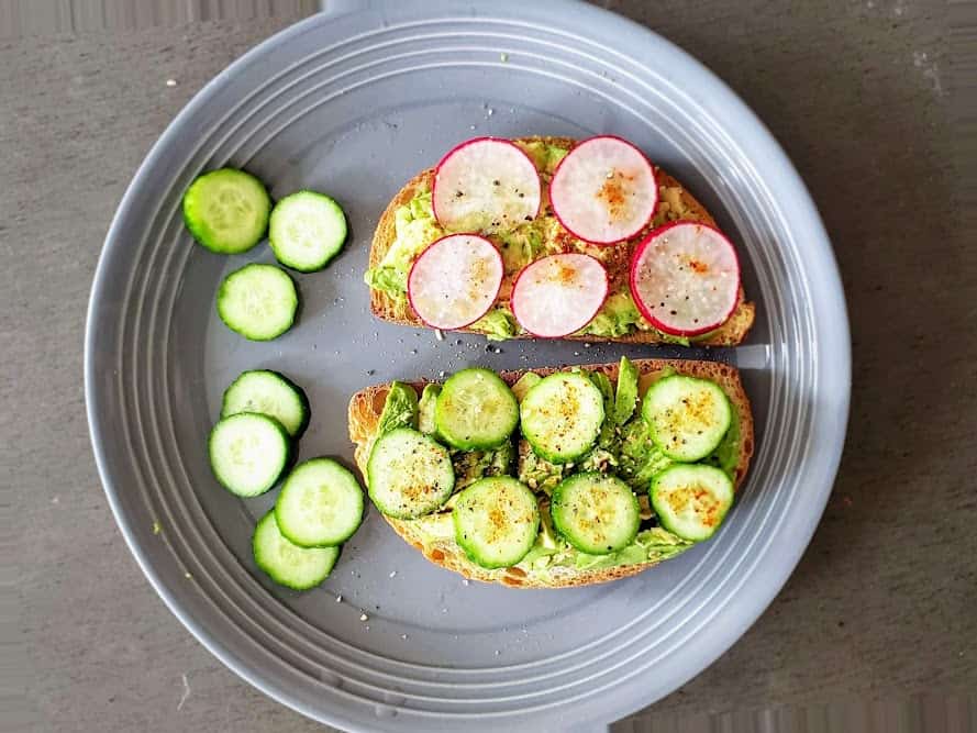 two avocado toasts with various slices of veggies served with red pepper seasoning. 