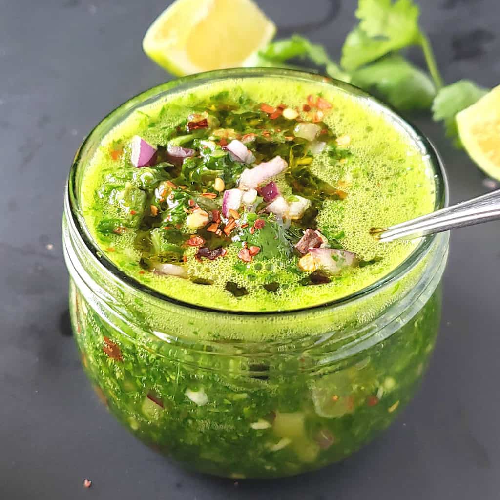 A glass jar full of bright green cilantro chimichurri sauce with red onion and red pepper flakes on top.