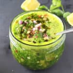 A glass jar full of bright green cilantro chimichurri sauce with red onion and red pepper flakes on top.