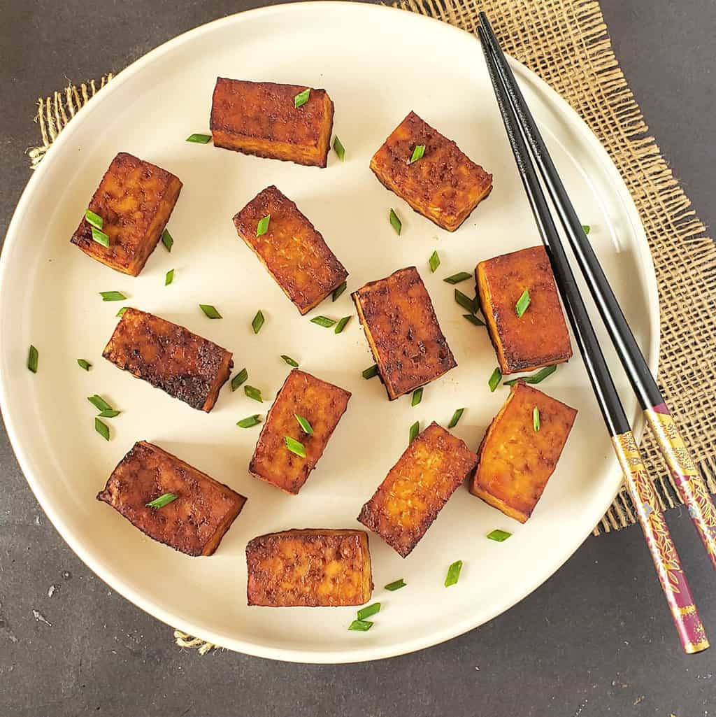 Smoky tofu served with garnishes on a white dinner plate with chop sticks..
