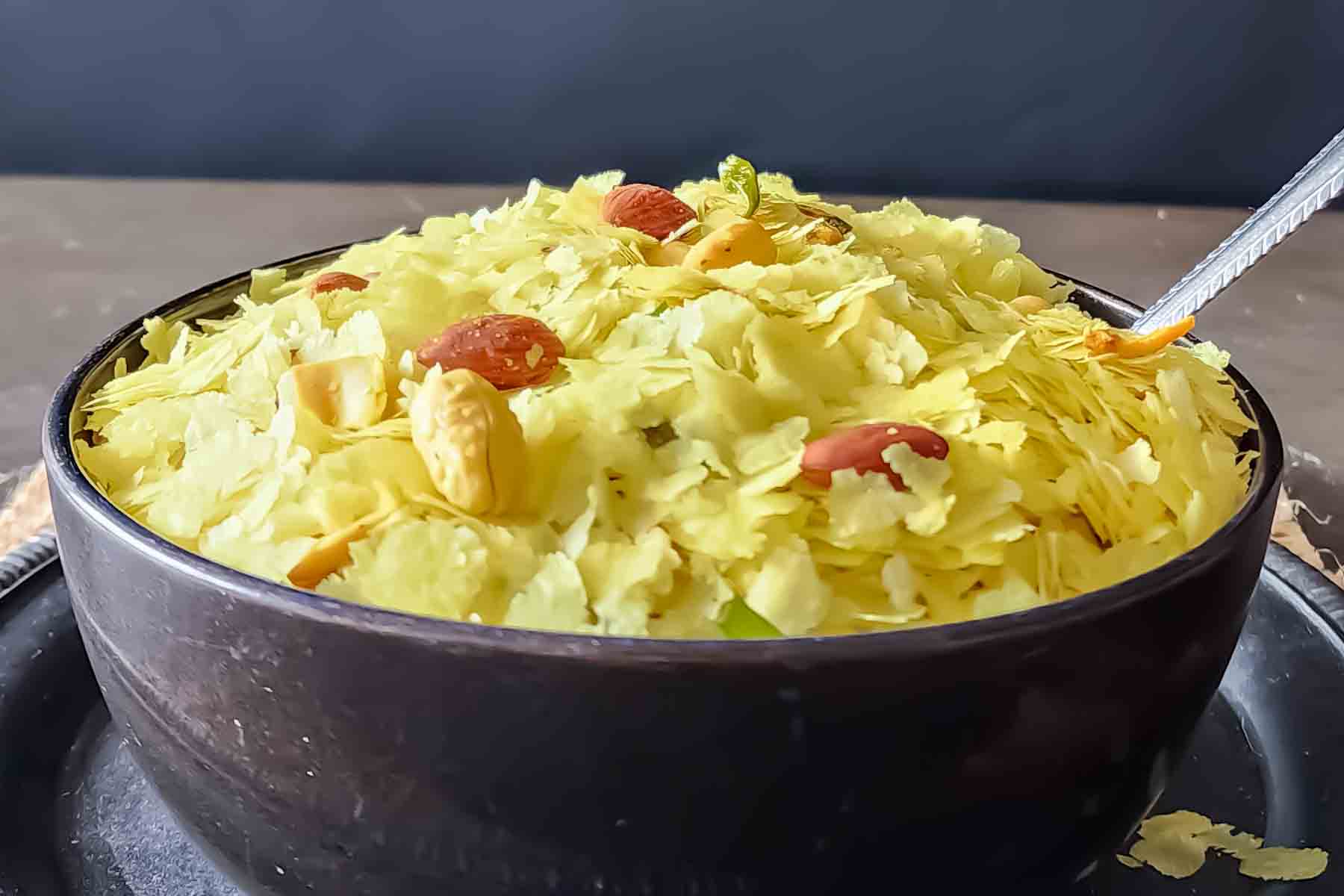A bowl with spoon filled with yellow golden hued indian snack chivda.
