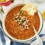 A soup bowl filled with homestyle lentil soup with vegetables, served with baguette, chopped onions, cilantro leaves and lemon wedges.